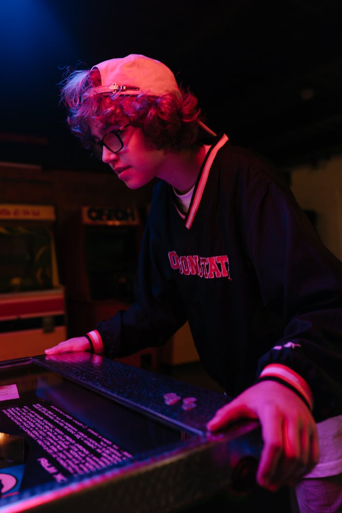 Teenage boy playing a retro pinball machine in a dimly lit arcade setting.